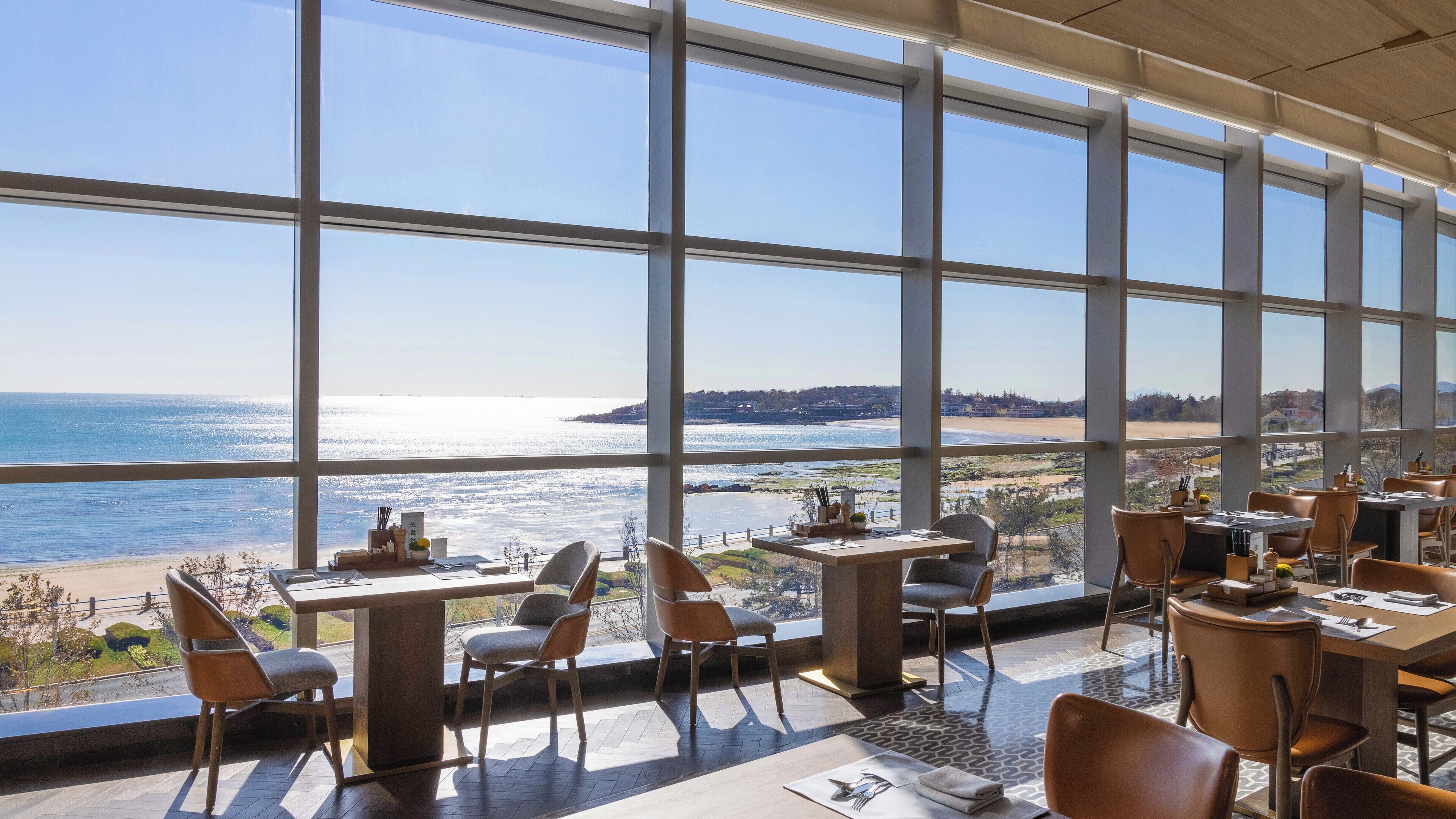 En restaurang med stora fönster som ger utsikt över stranden och havet. Bord och stolar är placerade ut för att skapa en trevlig matplats.