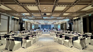 A conference room with rows of tables and chairs, a stage with a presentation screen, and a high ceiling with lighting fixtures.