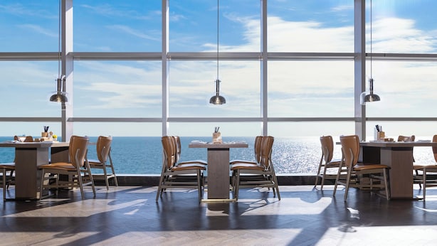 Een eetgedeelte met grote ramen met uitzicht op zee, voorzien van houten tafels en stoelen, en hanglampen.