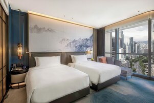 A modern hotel room with two beds, a city view, and a large artwork on the wall.