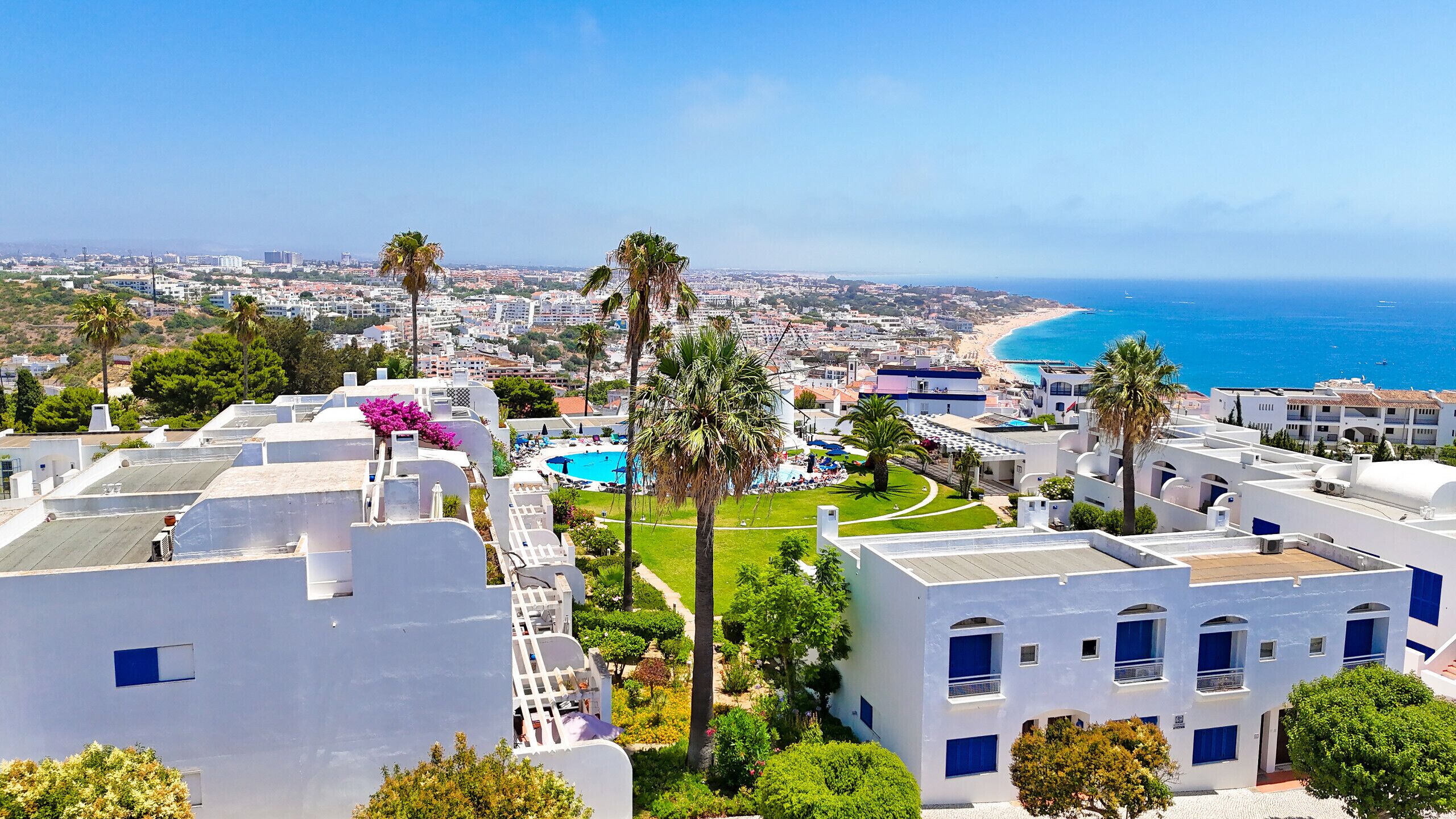 A coastal hotel with white buildings, a swimming pool, and a view of the sea and beach.