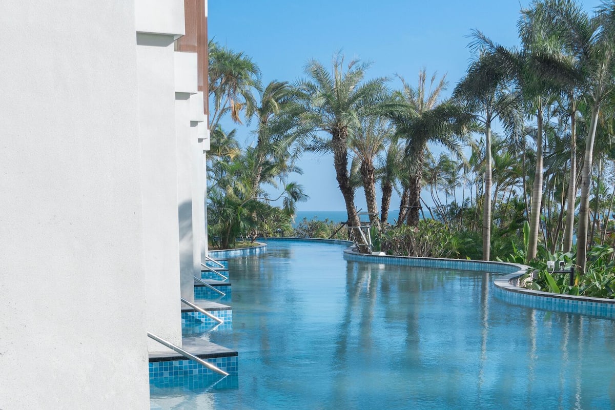 A swimming pool with clear blue water, surrounded by palm trees and a white building.
