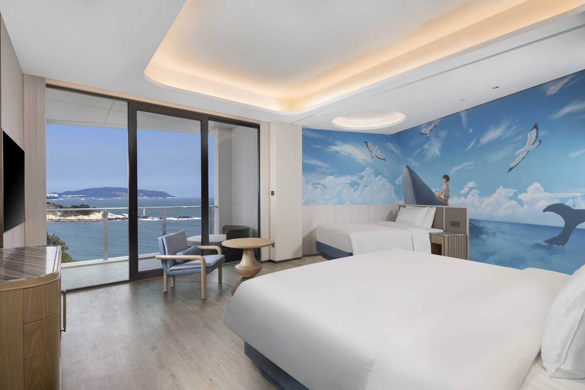 A modern hotel room with a large bed, a desk, and a view of the sea.