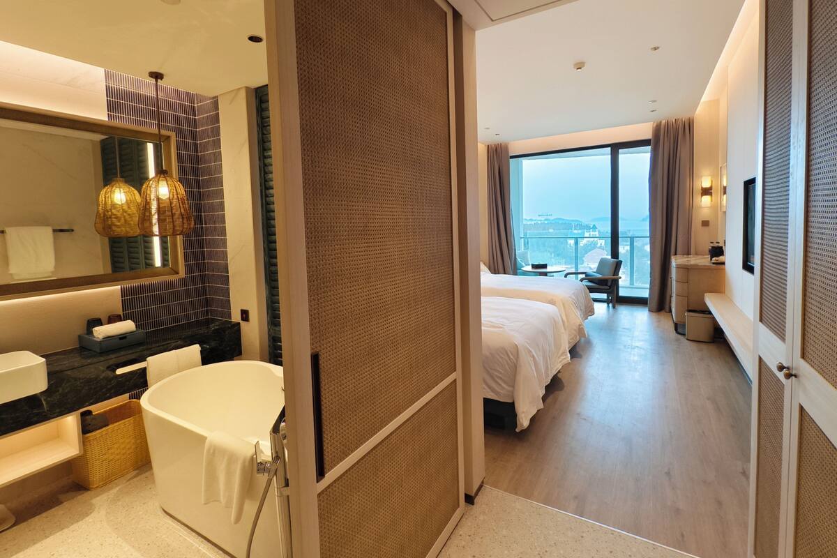 A modern hotel room with a large bed, a separate sitting area with a view, a bathroom with a bathtub, and a large mirror.
