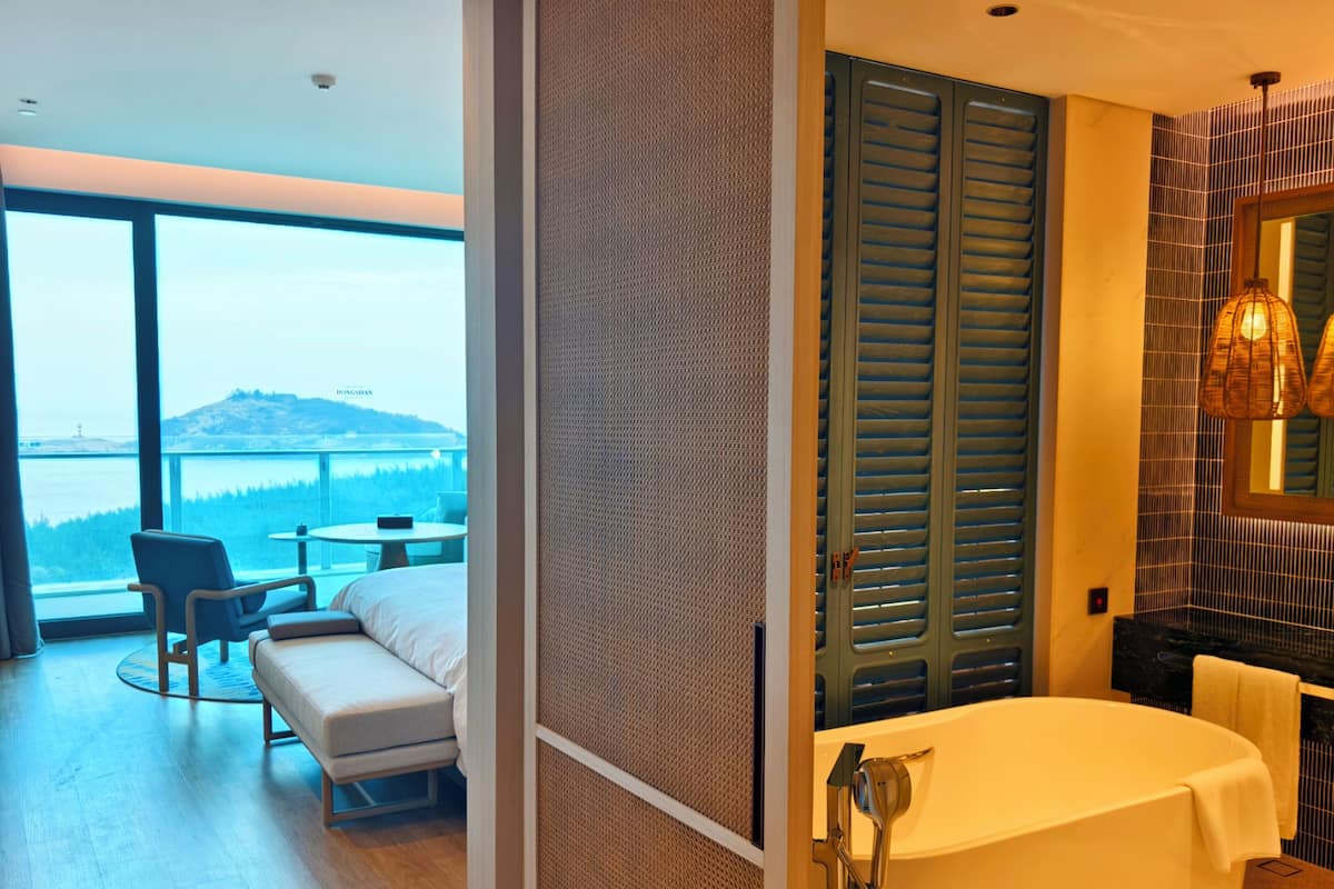 A modern hotel room with a large window offering a view of a body of water and a distant hill. The room includes a sofa, a small table, and a bathtub with a showerhead.