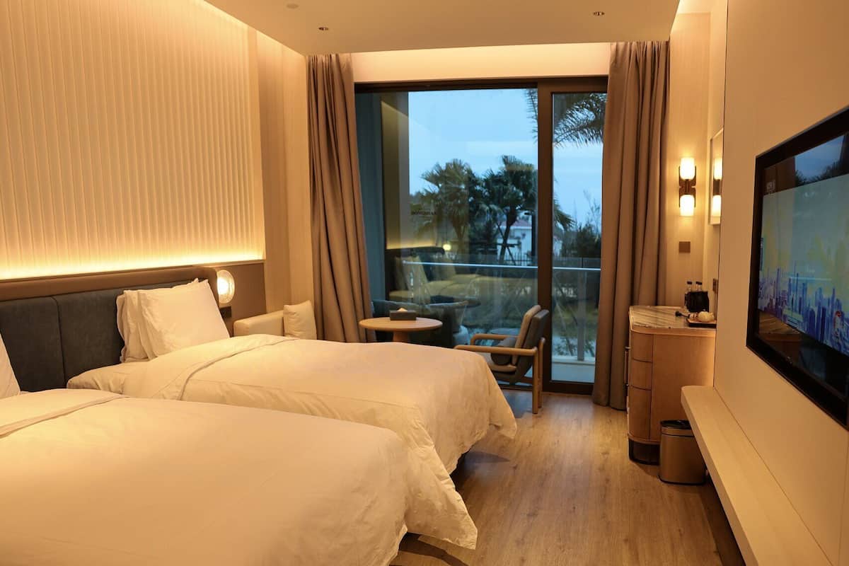 A hotel room with two beds, a small table, a chair, a TV, and a balcony with a view of palm trees.