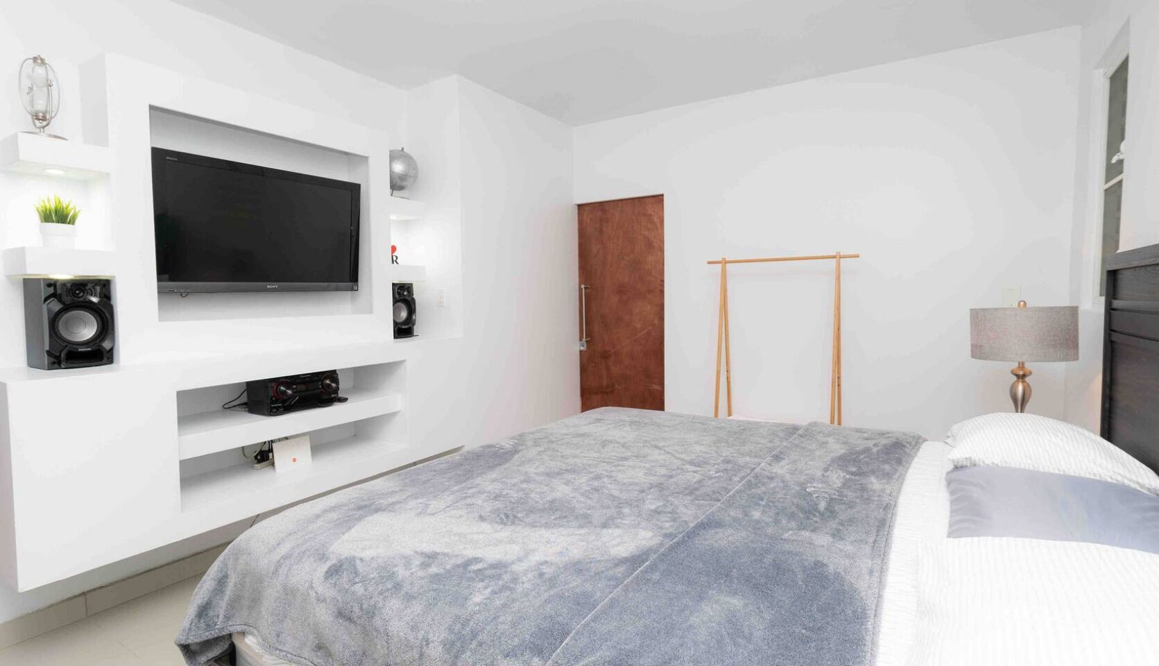 Ein modernes Schlafzimmer mit Flachbildfernseher, einem Holzschrank, einem Bett mit grauer Decke und einer Nachttischlampe.