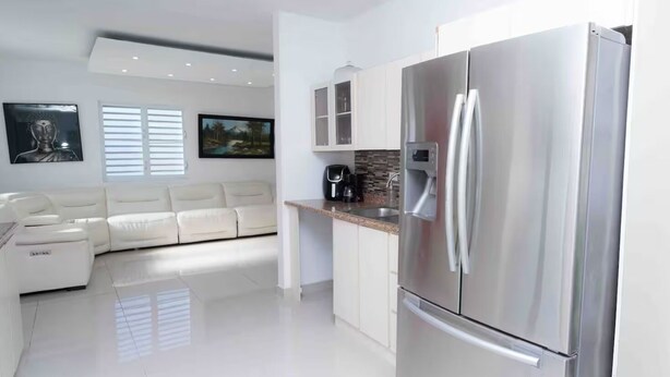 A modern kitchen with a stainless steel refrigerator, white cabinetry, and a white sofa in the adjacent living area. - Mystic River Retreat | 3BR/2BA•Pool - Jacuzzi - Beverly Hills, Guaynabo, Guaynabo, Puerto Rico (Guaynabo)