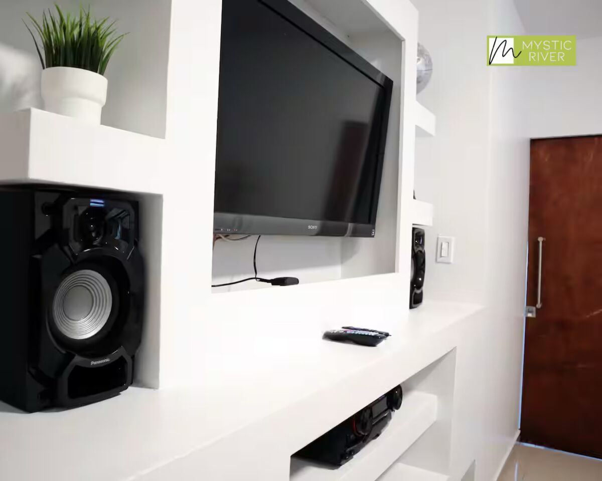 En moderne stue med et fladskærms-tv monteret på et hvidt tv-møbel, en sort højttaler og en fjernbetjening på møblet.