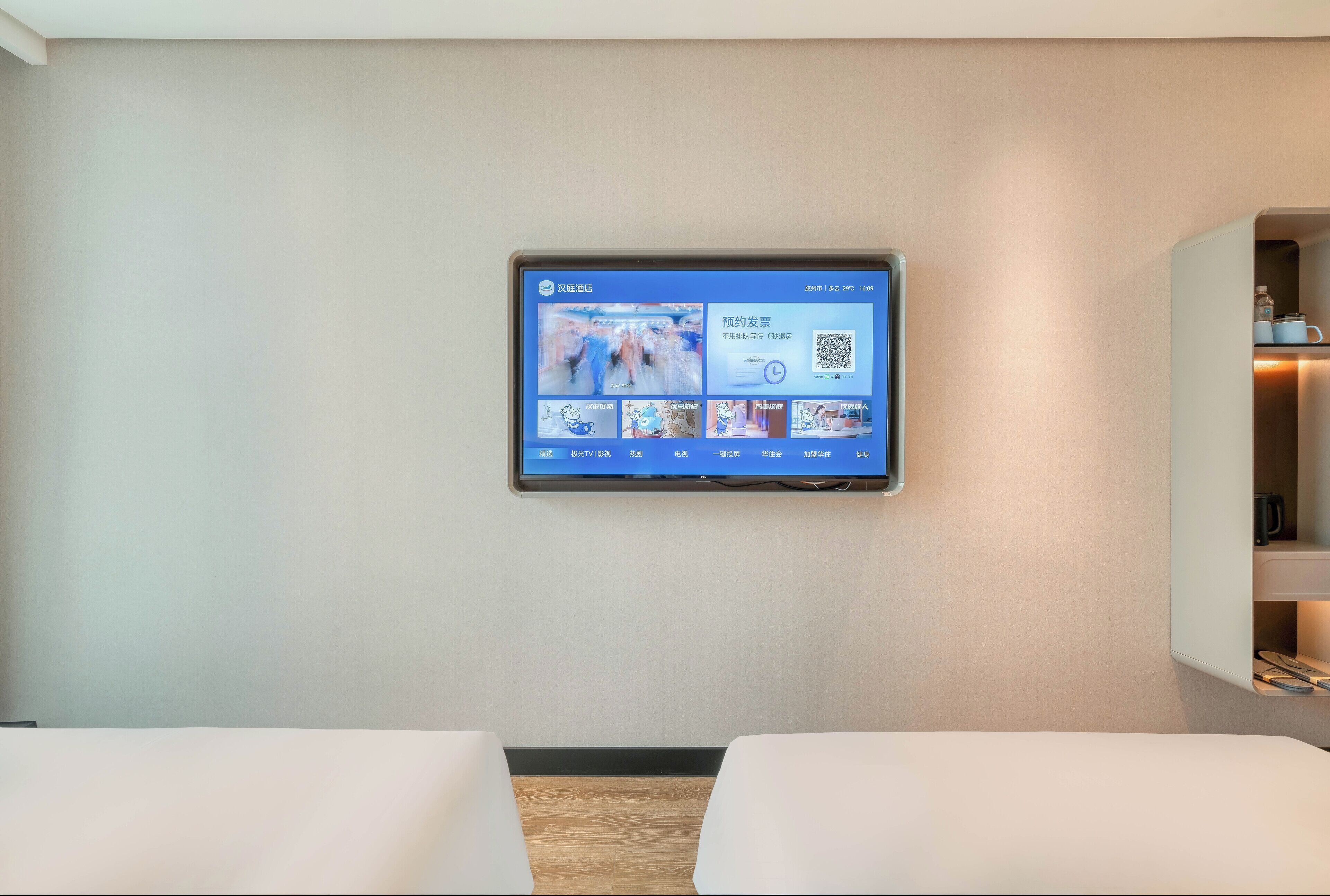 Habitació d'hotel amb televisió penjada que mostra un canal de notícies, un llit i una petita estant amb objectes.