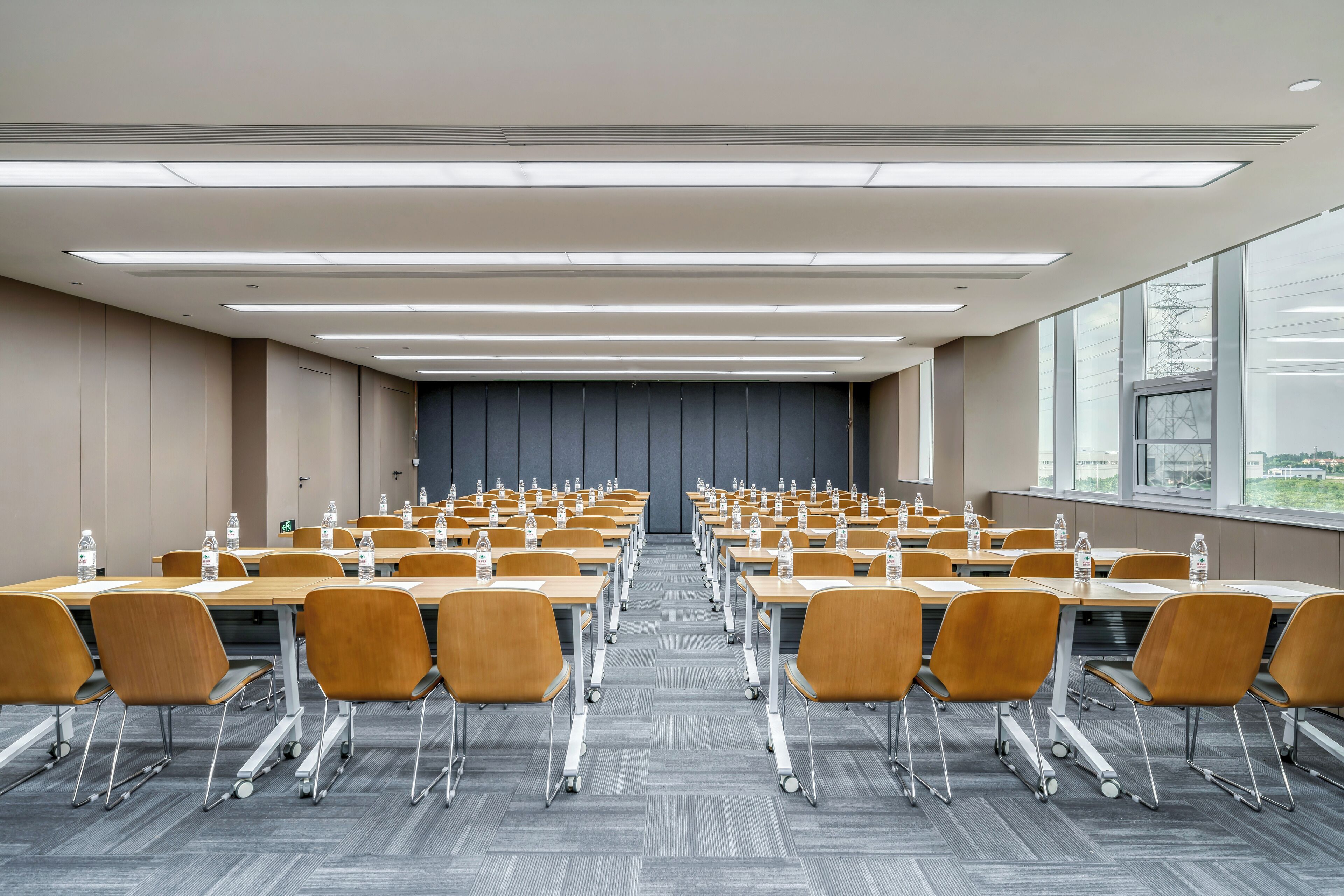 A conference room with rows of tables and chairs, equipped with water bottles, and large windows providing a view of the outside.
