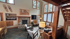 A spacious living room with a stone fireplace, a television mounted above it, and a large window with a view of snow-covered trees.