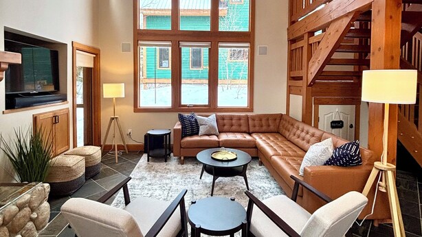 A spacious living room with a large sectional sofa, a coffee table, and a TV mounted on the wall. There are large windows providing natural light and a view of the outside.