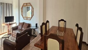 Ein geräumiges Wohnzimmer mit einem großen Esstisch aus Holz, sechs Stühlen, einem braunen Ledersofa, einem kleinen Beistelltisch und einem Fernseher auf einem Schrank.