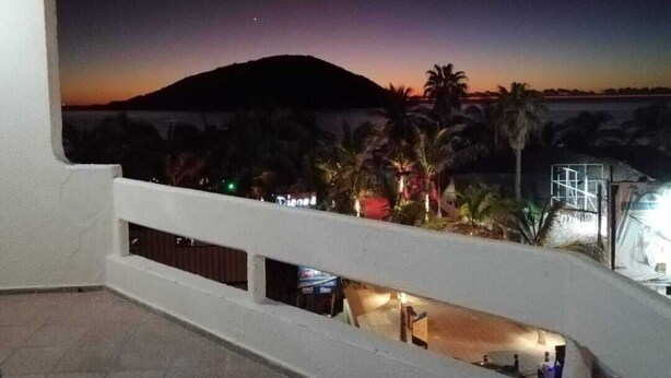 Ein Balkon mit Blick auf einen Sonnenuntergang über einer Hügelkette, Palmen und ein entferntes Gebäude.