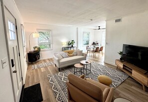 A modern living room with a sofa, coffee table, and TV stand. There is a dining area with chairs and a table. The room has hardwood flooring and patterned rugs. - Minutes to Masters! (Augusta)