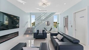 A modern living room with a flat-screen TV, black leather sofas, a glass coffee table, and a staircase with glass railings. - Marathon Sombrero Beach  5 min to Beach, Pool, 70' Dock, Elevator (Marathon)