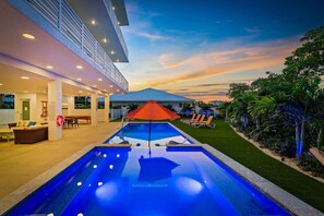 Uma área moderna com piscina ao ar livre, uma espreguiçadeira e guarda-sol, cercada por um gramado bem cuidado e plantas tropicais.