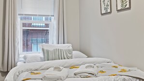 A neatly made bed with white bedding and a floral pattern, a window with blinds, and two framed botanical prints on the wall. - Prima Boston North End 2brm (Boston)