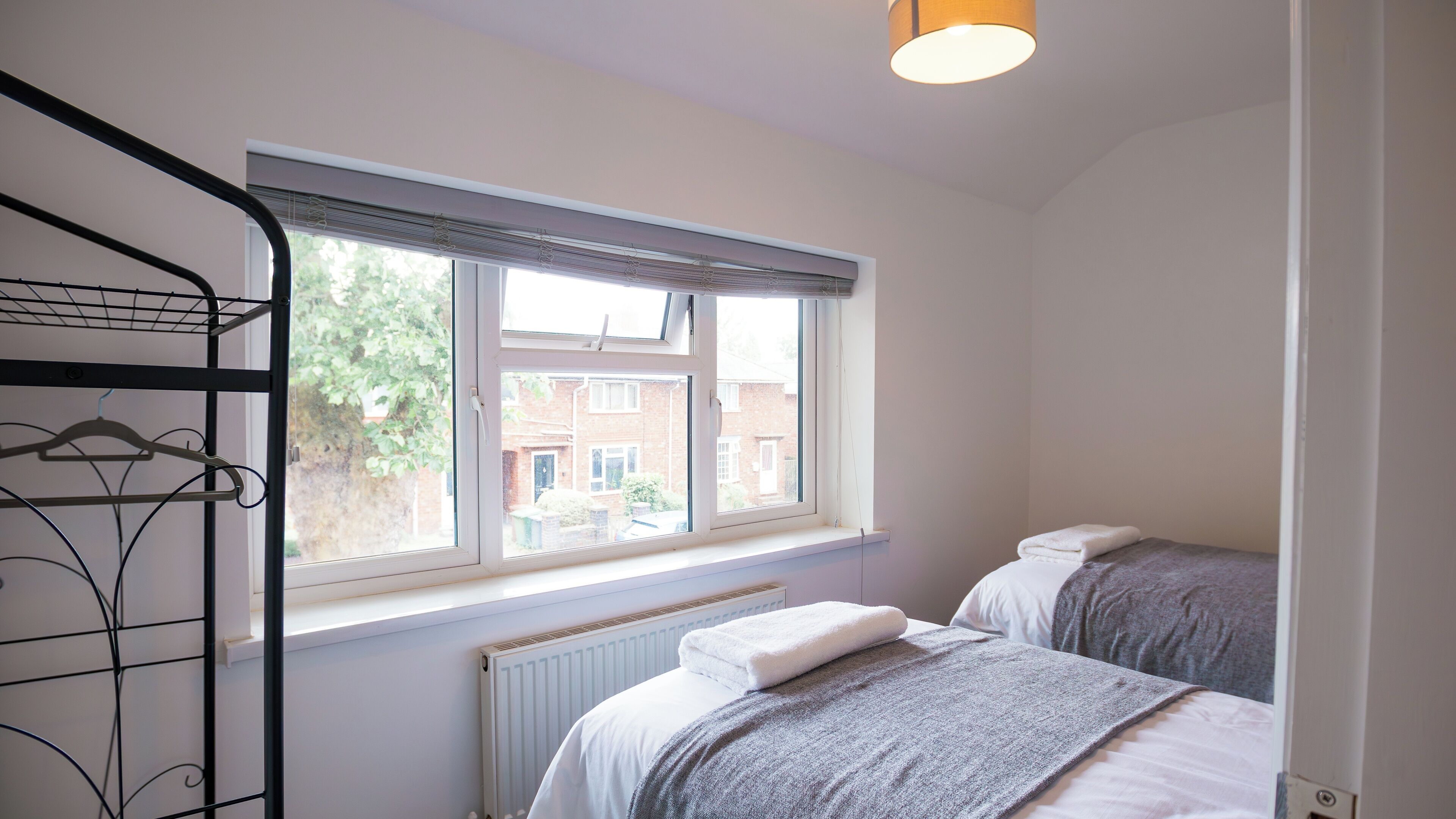 Een slaapkamer met twee bedden, een radiator, een raam met zonwering en een hanglamp.