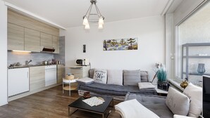 Una sala de estar moderna con un sofá, una mesa de centro y una mesita auxiliar. Hay una cocina con armarios y un microondas. Un cuadro cuelga en la pared, y una ventana ofrece vistas al exterior.