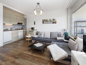 A modern living room with a sofa, coffee table, and a small side table. There is a kitchen area with cabinets and a microwave. A painting hangs on the wall, and a window provides a view of the outside. - Les Orzières 417 by Interhome (Crans-Montana)