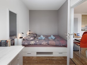 A compact bedroom with a bed, bedside table, lamp, and a view into the adjacent dining area.