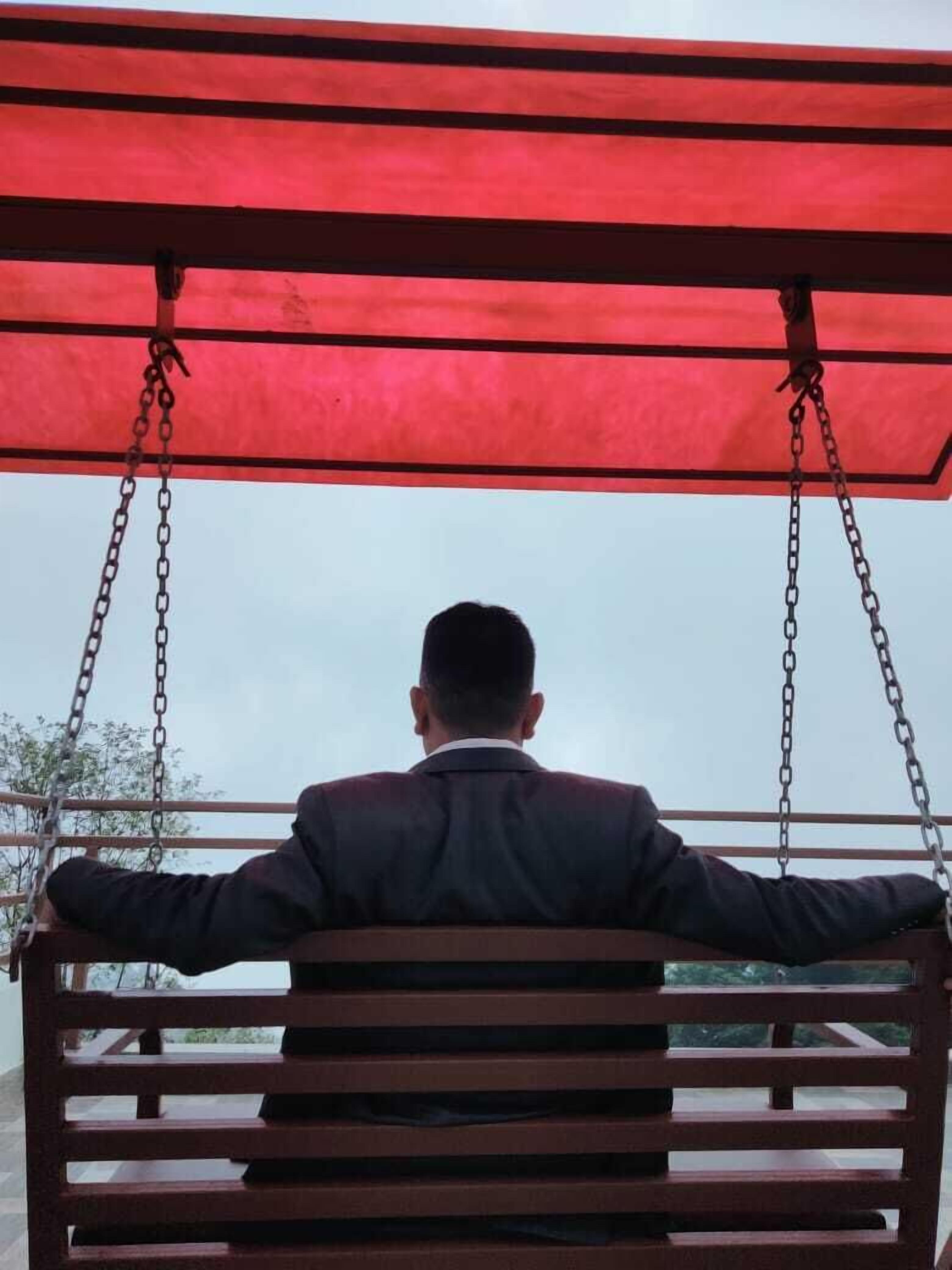 A man sitting on a swing with a red canopy, facing away from the camera.