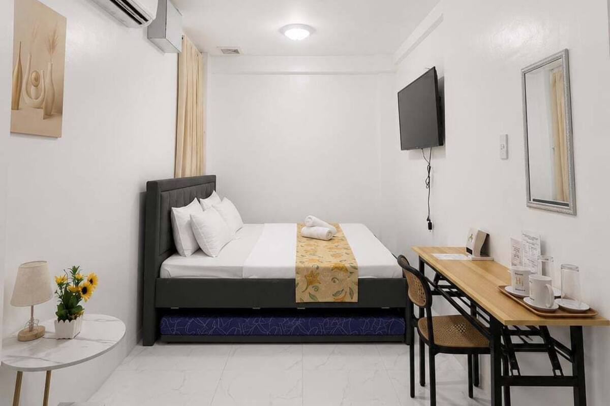 A hotel room with a bed, a desk, a chair, a TV, and an air conditioning unit.