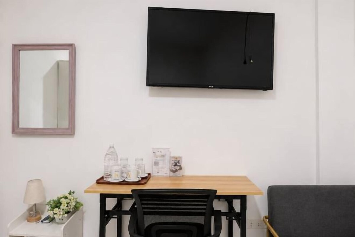 Une pièce dotée d’un téléviseur à écran plat fixé au mur, d’une table en bois avec des chaises, d’une petite table d’appoint avec une lampe et des fleurs, et d’un canapé gris.