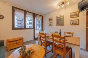 Un coin repas avec une table en bois, quatre chaises et un banc. Sur la table, on trouve quatre tasses, un gaufre et une bouteille de vin. La pièce est dotée d’une fenêtre avec des rideaux, d’une horloge au mur et de tableaux encadrés.