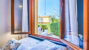 A bedroom with a large window, a bed with patterned bedding, and a view of a garden and building outside. - La Conchiglia by Interhome (Castiglioncello)