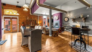 A spacious living room with a wooden floor, a dining area with a table and chairs, a fireplace, and a large window with a view. - The 1901 Loft - Heart of Old Colorado City, walking distance to everything (Colorado Springs)