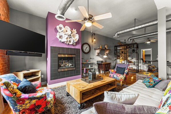 Un salón acogedor con chimenea, ventilador de techo y diversas opciones de asientos, incluyendo un sillón floral y un sofá.