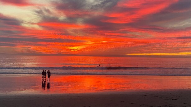 Uma praia com um pôr do sol, ondas e silhuetas de pessoas.