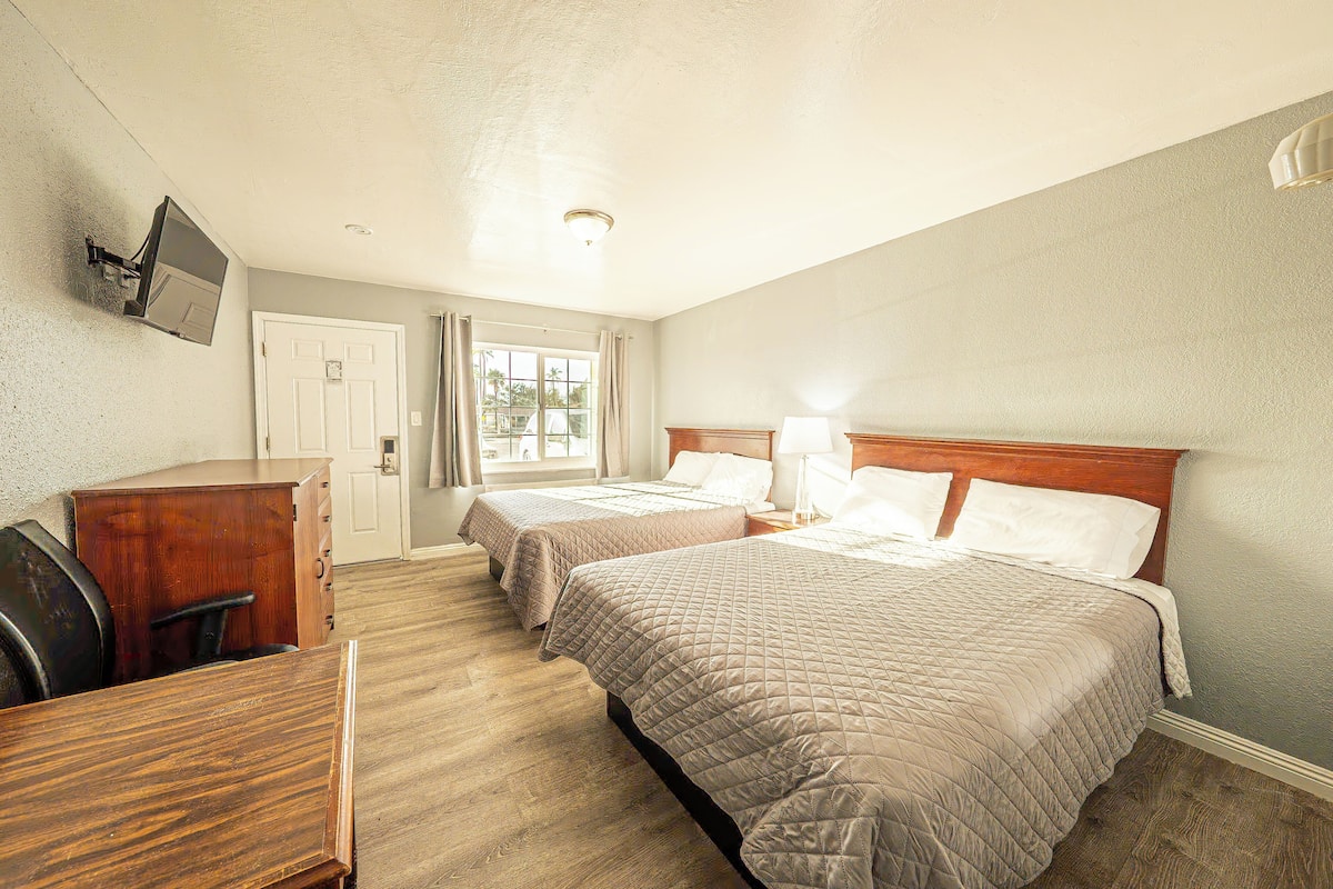 Habitación de hotel con dos camas, un escritorio, una silla, un televisor montado en la pared y un ventanal con cortinas.