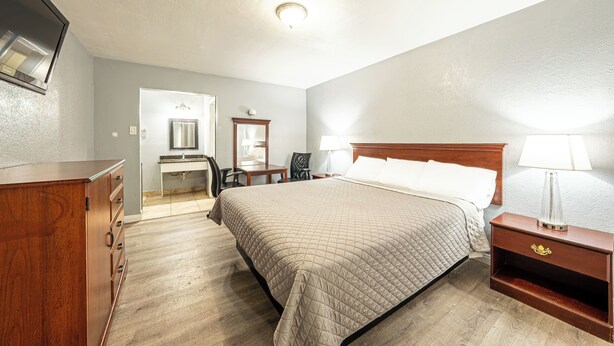 A hotel room with a bed, a wooden dresser, a desk with a chair, and a nightstand with a lamp. - Villa Motel (Santa Maria)