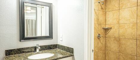 A bathroom with a granite countertop sink, a mirror, and a shower with beige tiles.