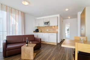 Una sala de estar moderna con un sofá marrón, una mesa de centro de madera y una cocina con armarios blancos y un microondas.