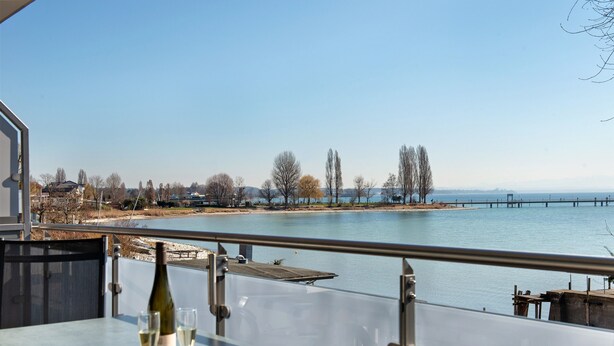 A table with a bottle and glasses, overlooking a body of water with a dock and trees. - Apartment 'Typ A2 2 Obergeschoss Nummer 13' with Shared Terrace, Shared Garden and Wi-Fi (Immenstaad am Bodensee)