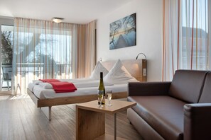 Habitación de hotel moderna con una cama, una mesita de noche de madera, una mesa de vidrio con una botella y un vaso, un sofá de cuero marrón y ventanas amplias con cortinas.
