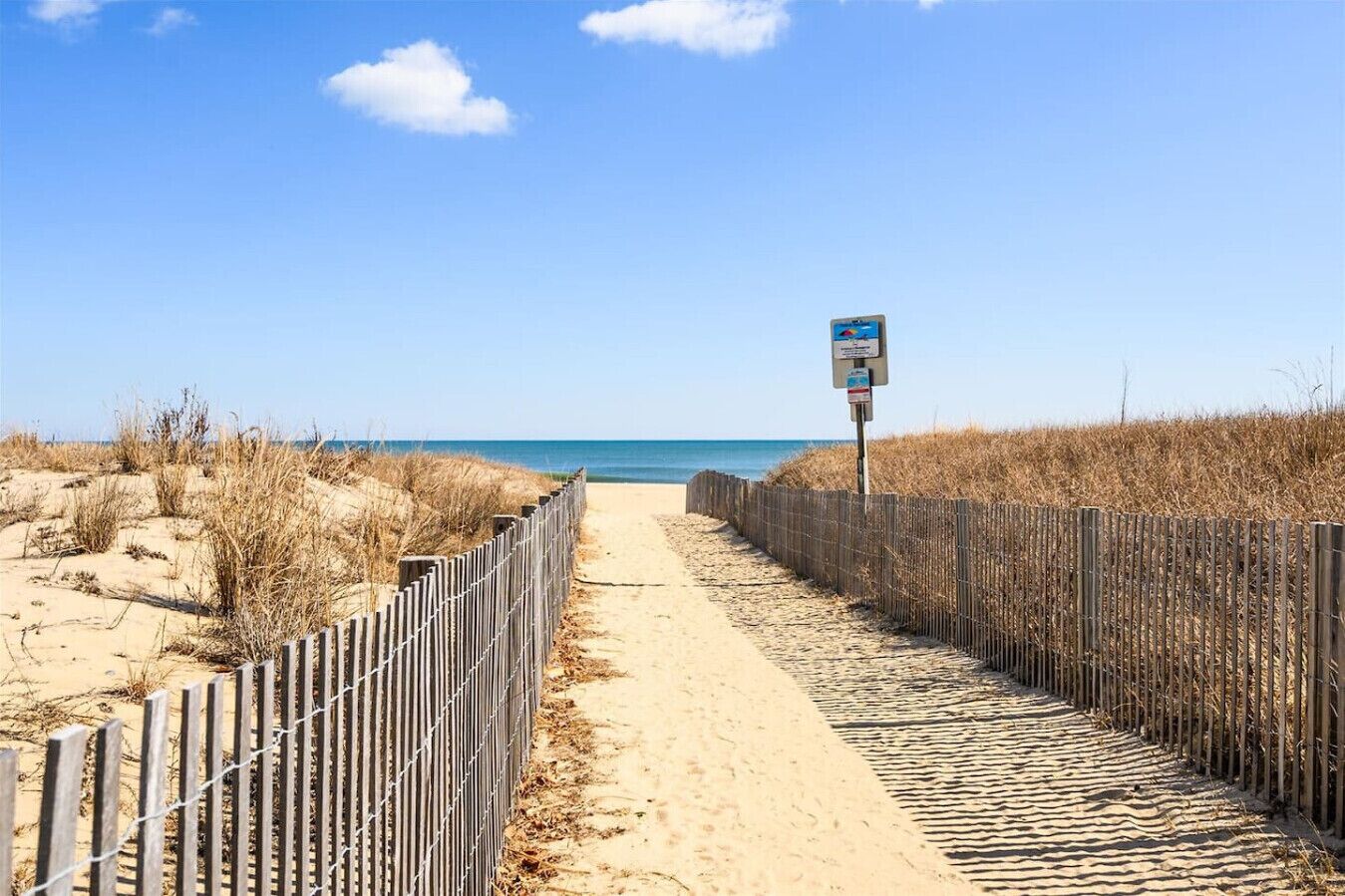 Un sentier de plage sablonneux, bordé d’une clôture en bois d’un côté et d’un panneau indicateur visible au loin.