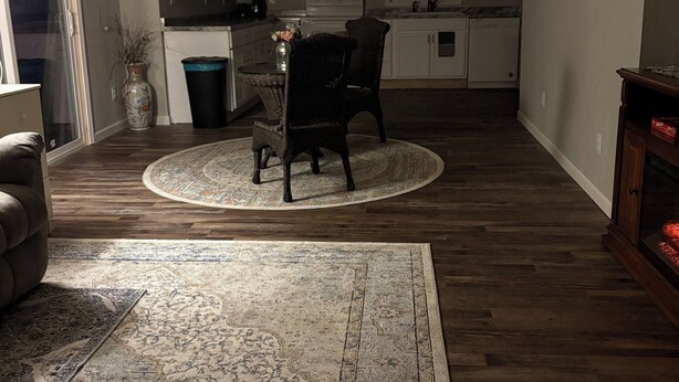 Un salon avec un tapis rond, un canapé de couleur sombre, un coin repas avec une chaise en osier et, au fond, une cuisine.