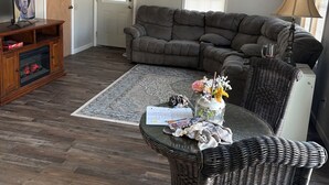Un salon comprenant un canapé d’angle, un fauteuil en osier, une petite table avec des papiers et un vase, ainsi qu’un téléviseur posé sur un meuble en bois.