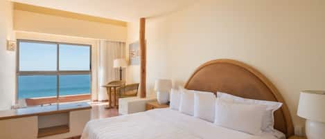 A hotel room with a large bed, a ceiling fan, a view of the ocean, a wooden nightstand, and a window with curtains.