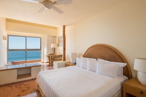Ein Hotelzimmer mit einem großen Bett, einem Deckenventilator, Blick aufs Meer, einem Holznachttisch und einem Fenster mit Vorhängen.