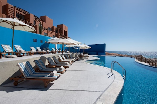 Ein Poolbereich mit Liegestühlen und Sonnenschirmen, mit Blick auf das klare Meer, und ein Gebäude mit blau-brauner Fassade.
