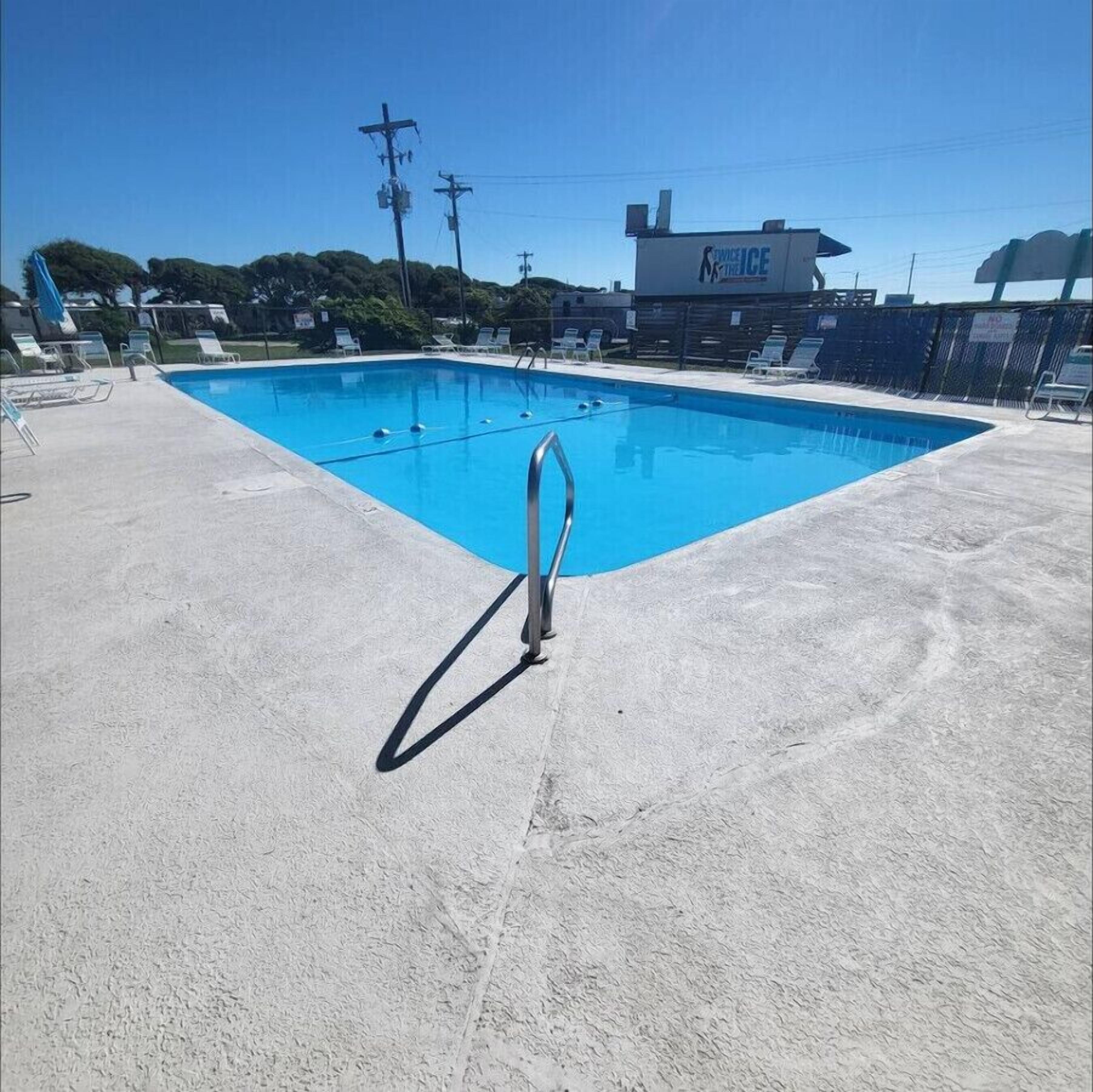 Una piscina con escalera metálica, rodeada de sillas de descanso y un suelo de hormigón.
