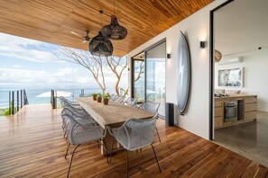 Un espace repas moderne avec une table et des chaises en bois, une vue sur l’océan et un coin cuisine doté de meubles en bois.