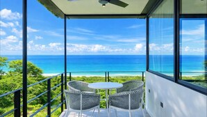 Ein Balkon mit Tisch und zwei Stühlen, mit Blick auf ein klares blaues Meer und üppiges Grün.